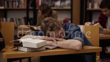年轻的欧洲学生在大学图书馆或<strong>班级</strong>睡在桌子上，上面有一堆书。 同学和书架